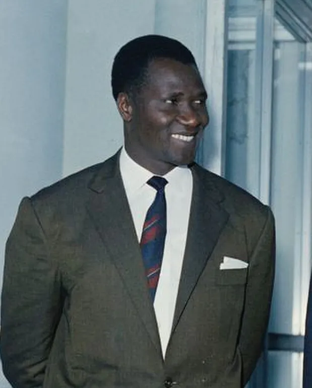 Ahmed Sékou Touré, sonriente, con un traje oscuro, camisa blanca y corbata a rayas, de pie frente a una ventana.